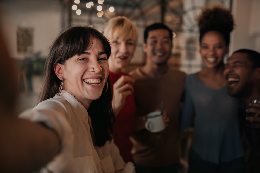 Laughing group of diverse young friends talking a selfie together while having drinks in a bar in the evening